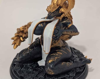 May include: A fantasy figurine of a black and gold dragon-like creature with large curved horns, kneeling on a cracked circular base. The figure features textured scales, gold accents, and a long white cloth draped over the chest and torso.