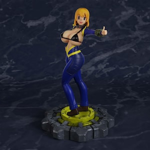 May include: A collectible figurine of a character with short blonde hair, wearing a black bikini top and a blue and yellow jumpsuit, standing on a circular gear-shaped base. The character is smiling and giving a thumbs-up gesture with one hand.