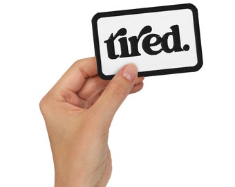 May include: A white patch with a black border and the word "tired." in black lettering. The patch is being held by a hand.