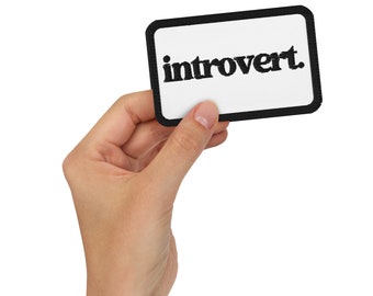 May include: A white rectangular patch with a black border, held in a hand. The word "introvert" is printed in black, stylized font across the center of the patch. The background is plain white.