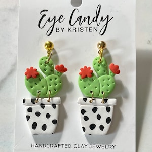 May include: A pair of green cactus earrings with red flowers and black and white polka dot pots. The earrings are handmade and say "Eye Candy by Kristen" and "Handcrafted Clay Jewelry".