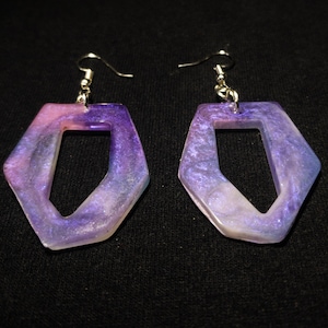 Peut inclure: Une paire de boucles d'oreilles en résine violette et blanche avec une forme géométrique et un crochet argenté.