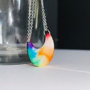 Può includere: Una collana con catena d'argento con un ciondolo a forma di mezzaluna arcobaleno. Il ciondolo è in resina e presenta un motivo a spirale bianco, arancione, giallo, verde, blu e viola.
