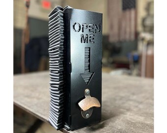 May include: Black metal bottle opener with a built-in bottle holder. The opener has a cutout that says "OPEN ME" with a downward arrow. The bottle opener is on a gray surface.