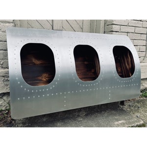 May include: A silver metal airplane fuselage section with three oval-shaped windows. The windows are framed in black and have a wood-like interior. The fuselage is covered in rivets.