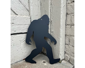 May include: Black metal silhouette of Bigfoot walking, cut out from a sheet of metal. The silhouette is placed against a white brick wall.