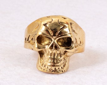 May include: A gold-coloured skull ring with intricate details. The skull is depicted with a detailed face and a textured surface.
