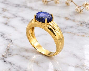 May include: A gold ring featuring a polished, smooth band and a central oval-shaped lapis lazuli gemstone. The deep blue stone is secured by four prongs, showcasing natural gold-colored flecks within the surface of the dark blue mineral.
