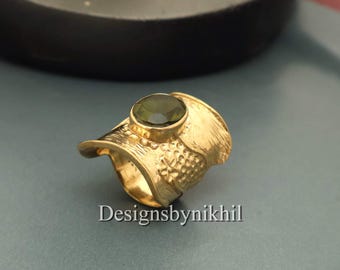 Può includere: Un anello color oro con una gemma ovale verde oliva. L'anello ha una fascia strutturata e un design decorativo sul lato. Il testo "Designsbynikhil" è visibile sulla superficie.