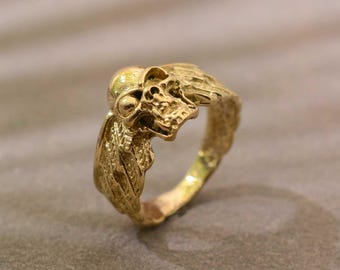 May include: A gold ring with a skull design. The skull is the central feature, with detailed facial features. The ring has a textured band and a gothic or biker style, with a vintage appearance.