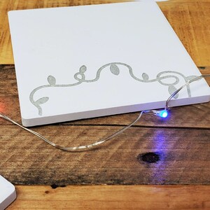 以下が含まれることがあります： 上部に蔦のデザインが施された白い正方形の木製看板2枚。看板は木製の表面に置かれ、赤、青、黄色の電球が付いた白い電飾が飾られています。