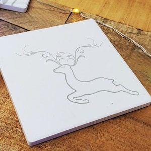 以下が含まれることがあります： 白いセラミックコースターに、グレーの輪郭で描かれた鹿のシルエット。鹿は右に向かって走っている。