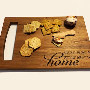 May include: A wooden serving board with a handle, engraved with the words "home" and coordinates: 30° 24' 49.86" N 87° 12' 58.5" W. The board has crackers and a small round cheese on it.
