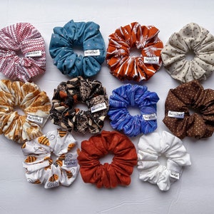 May include: A collection of 11 colorful hair scrunchies with various patterns, including stripes, polka dots, and floral designs. Each scrunchie has a small tag that reads "Made by Heather".