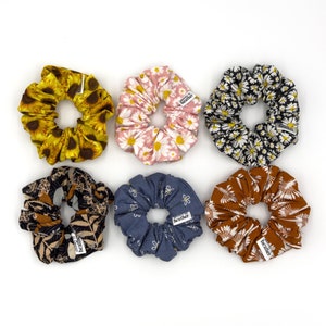 May include: Six colorful scrunchies with floral and abstract patterns. The scrunchies are made of fabric and have a small tag with the word "Heather" on them.