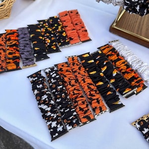 May include: A collection of fabric scrunchie watch bands featuring various Halloween-themed patterns including orange pumpkins, white ghosts, black bats, and candy corn on black, orange, and purple backgrounds with metal connectors.