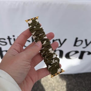 May include: A scrunchie-style watch band featuring an olive green and white cow print pattern. The fabric band is gathered with elastic and includes gold-toned metal connectors at both ends for attachment to a smartwatch device.