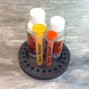 May include: A circular gray plastic organizer stand featuring twenty-four numbered holes around the perimeter. Two medicine bottles with white caps and two oral syringes, one yellow labeled Infants' Tylenol and one orange labeled Infants' Motrin, are placed inside the stand.