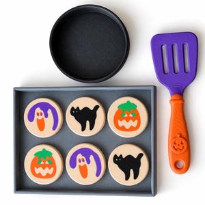 May include: A set of six round, light-colored wooden discs featuring printed Halloween designs of ghosts, black cats, and jack-o'-lanterns, arranged in a gray rectangular tray, accompanied by a purple and orange spatula with a pumpkin motif.