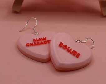 Può includere: Orecchini rosa a forma di cuore con le parole "MAIN CHARACTER" e "BOUJEE" in lettere rosse. Gli orecchini hanno ganci color argento. Lo sfondo è di colore rosa.