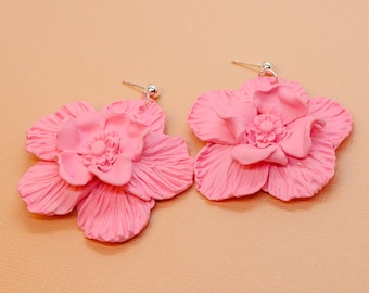 Può includere: Un paio di orecchini a forma di fiore rosa. Ogni orecchino presenta un design floreale dettagliato e stratificato con una superficie strutturata. Gli orecchini hanno perni color argento e piccoli accenti a sfera. Lo sfondo è di colore pesca chiaro.