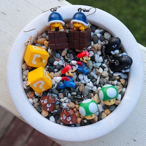 Pu&ograve; includere: Una collezione di orecchini originali con personaggi e oggetti della serie di videogiochi Super Mario. Gli orecchini includono blocchi gialli con punti interrogativi, figure di Mario rosso e blu e disegni di funghi verdi. Gli orecchini sono esposti in una ciotola bianca piena di piccole pietre.