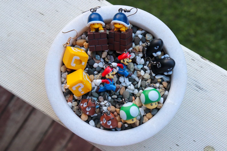 Pu&ograve; includere: Una collezione di orecchini originali con personaggi e oggetti della serie di videogiochi Super Mario. Gli orecchini includono blocchi gialli con punti interrogativi, figure di Mario rosso e blu e disegni di funghi verdi. Gli orecchini sono esposti in una ciotola bianca piena di piccole pietre.