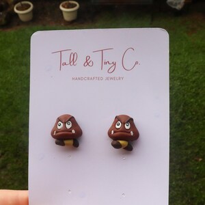 May include: A pair of brown Goomba earrings with white eyes and yellow accents. The earrings are displayed on a white card with the text "Tall & Tiny Co. HANDCRAFTED JEWELRY". The background is a blurred outdoor scene.