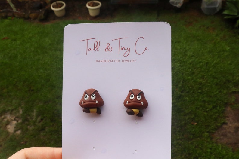 May include: A pair of brown Goomba earrings with white eyes and yellow accents. The earrings are displayed on a white card with the text "Tall & Tiny Co. HANDCRAFTED JEWELRY". The background is a blurred outdoor scene.