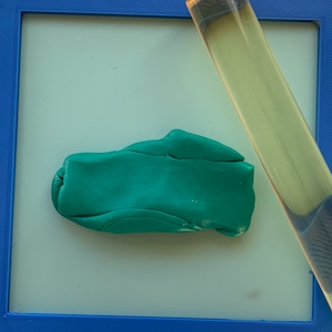 May include: A piece of green modeling clay shaped into a rectangular form, positioned on a light blue surface within a blue frame. A clear rolling pin rests diagonally across the frame, next to the clay. The scene is brightly lit.