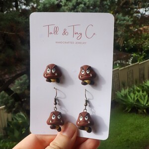 May include: Handcrafted jewelry featuring two pairs of brown Goomba earrings and studs. The earrings and studs are displayed on a white card with the text "Tall & Tiny Co. HANDCRAFTED JEWELRY". The Goombas have angry expressions.
