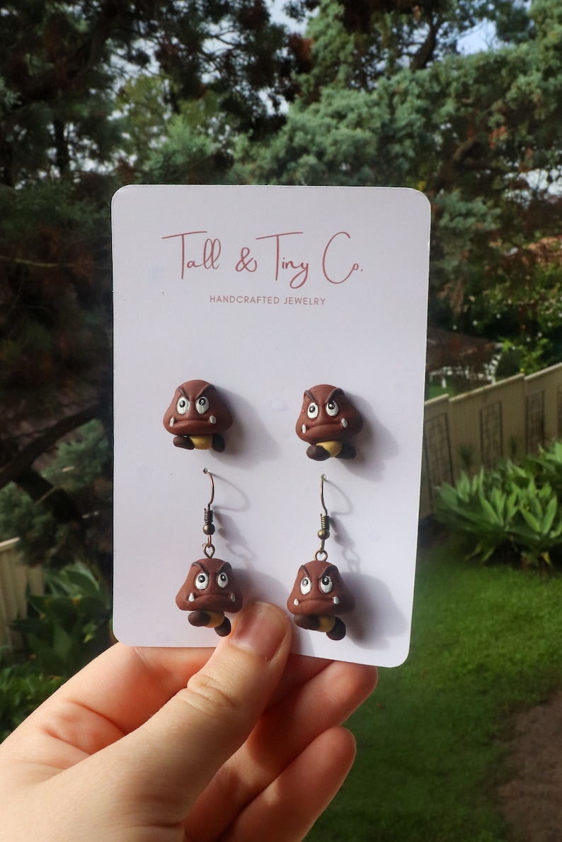 May include: Handcrafted jewelry featuring two pairs of brown Goomba earrings and studs. The earrings and studs are displayed on a white card with the text "Tall & Tiny Co. HANDCRAFTED JEWELRY". The Goombas have angry expressions.