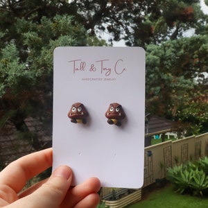 May include: A pair of brown Goomba earrings on a white card with the text "Tall & Tiny Co. HANDCRAFTED JEWELRY". The earrings are shaped like the video game character Goomba, with a brown body, white eyes, and a yellow base.