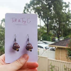May include: Goomba-shaped earrings made of brown clay with black metal hooks. The earrings are on a white card with the text "Tall & Tiny Co. HANDCRAFTED JEWELRY". The earrings are a playful accessory.