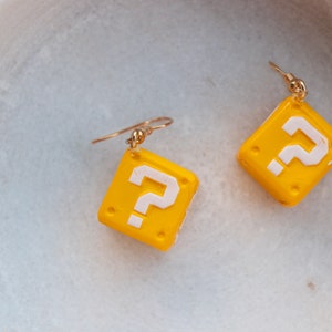 Pu&ograve; includere: Un paio di orecchini gialli e bianchi a forma di punto interrogativo. Gli orecchini sono di forma quadrata con un gancio color oro. Il punto interrogativo &egrave; bianco e lo sfondo &egrave; giallo. Gli orecchini sono probabilmente fatti di argilla polimerica.