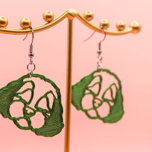 May include: A pair of green, open-design earrings shaped like a panda's face, hanging from silver hooks. The earrings are displayed on a gold-colored jewelry stand against a soft pink background. The panda design is a repeating pattern.