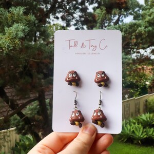 May include: Handcrafted jewelry set featuring Goomba earrings and studs. The earrings and studs are brown with a yellow underside and white eyes. The card reads "Tall & Tiny Co. HANDCRAFTED JEWELRY". The jewelry is displayed against a backdrop of green foliage.