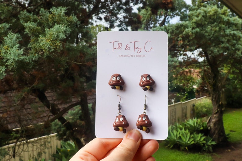 May include: Handcrafted jewelry set featuring Goomba earrings and studs. The earrings and studs are brown with a yellow underside and white eyes. The card reads "Tall & Tiny Co. HANDCRAFTED JEWELRY". The jewelry is displayed against a backdrop of green foliage.