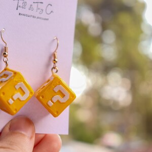 Pu&ograve; includere: Orecchini a forma di cubo giallo con punti interrogativi bianchi, ispirati a un videogioco. Gli orecchini sono appesi a ganci color oro. Il testo "Tall & Tiny Co. HANDCRAFTED JEWELLERY" &egrave; stampato sulla carta.