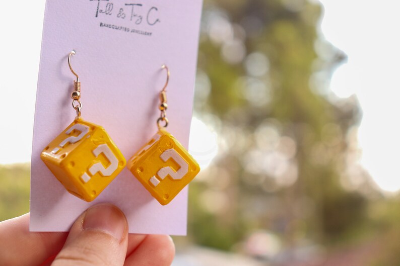 Pu&ograve; includere: Orecchini a forma di cubo giallo con punti interrogativi bianchi, ispirati a un videogioco. Gli orecchini sono appesi a ganci color oro. Il testo "Tall & Tiny Co. HANDCRAFTED JEWELLERY" &egrave; stampato sulla carta.