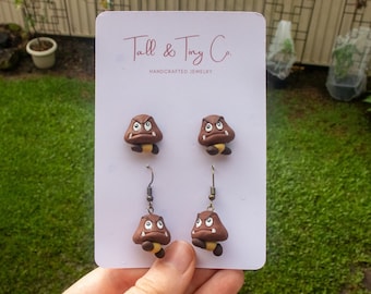 Può includere: Un paio di orecchini a lobo e un paio di orecchini pendenti con personaggi marroni a forma di fungo con espressioni arrabbiate. Gli orecchini sono esposti su una carta bianca con il testo "Tall & Tiny Co. HANDCRAFTED JEWELRY".
