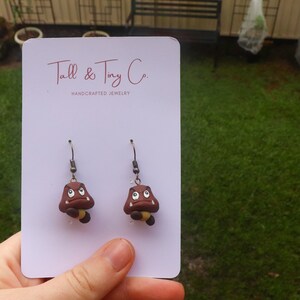 May include: A pair of handcrafted earrings featuring brown Goomba characters with white eyes and yellow accents. The earrings are displayed on a white card with the text "Tall & Tiny Co. HANDCRAFTED JEWELRY".