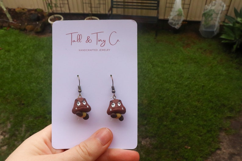 May include: A pair of handcrafted earrings featuring brown Goomba characters with white eyes and yellow accents. The earrings are displayed on a white card with the text "Tall & Tiny Co. HANDCRAFTED JEWELRY".