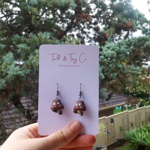 May include: A pair of handmade earrings featuring brown and tan Goomba characters from the Super Mario video game series. The earrings are attached to a white card with the text "Tall & Tiny Co. HANDCRAFTED JEWELRY".