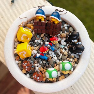 May include: A collection of novelty earrings featuring characters and elements from the Super Mario video game series. The earrings include yellow question mark blocks, mushrooms, and characters like Mario and Bullet Bills.