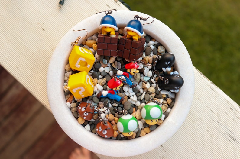 May include: A collection of novelty earrings featuring characters and elements from the Super Mario video game series. The earrings include yellow question mark blocks, mushrooms, and characters like Mario and Bullet Bills.