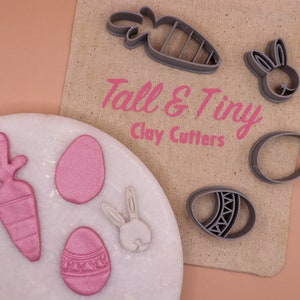 Peut inclure: Un assortiment d'emporte-pièces et de créations en argile sur le thème de Pâques. Les emporte-pièces sont en plastique gris en forme de carotte, de lapin et d'œufs. Les créations en argile sont roses et blanches. Le texte "Tall & Tiny Clay Cutters" est imprimé sur un sac en toile de jute.