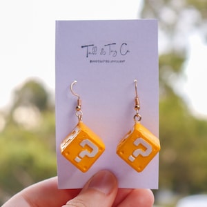 Pu&ograve; includere: Un paio di orecchini fatti a mano con un design a cubo giallo e un punto interrogativo bianco su ogni lato. Gli orecchini hanno ganci color oro e sono esposti su una carta bianca con il testo "Tall & Tiny Co. HANDCRAFTED JEWELLERY".