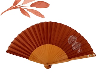Puede incluir: Un abanico de mano marrón con mango de madera y un diseño floral blanco. El abanico está cerrado y tiene un patrón decorativo en la parte posterior.