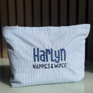 May include: A blue and white striped seersucker pouch. The pouch has the name "Harlyn" embroidered in dark blue, with the words "Nappies & Wipes" underneath. Ideal for storing baby nappies and wipes.